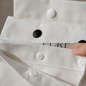 May include: Close-up of white fabric with black and white buttons. The fabric appears to be a shirt collar or similar garment detail. The buttons are round and sewn onto the fabric. The image is well-lit, highlighting the texture of the fabric.