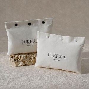 May include: Two off-white fabric pouches with black button closures and the word "PUREZA" printed in black. The word "PACKAGE" is printed below "PUREZA". One pouch is on a textured stone.