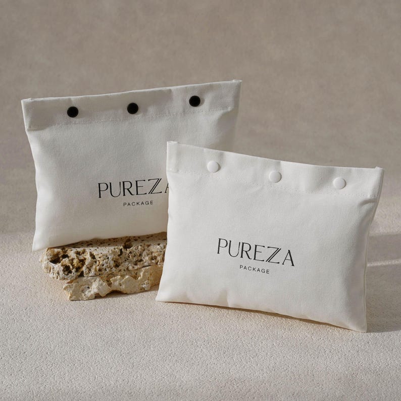 May include: Two off-white fabric pouches with black button closures and the word "PUREZA" printed in black. The word "PACKAGE" is printed below "PUREZA". One pouch is on a textured stone.