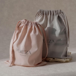 May include: Two drawstring bags, one blush pink and one light gray, are displayed. The pink bag has the word "PUREZA PACKAGE" printed in black. The gray bag has the word "PUREZA" printed in black. Both bags are made of a soft fabric.