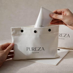 May include: A white canvas bag with the text "PUREZA PACKAGE" in black. The bag is open, and a white tube is being placed inside. The bag has three black snap closures at the top.