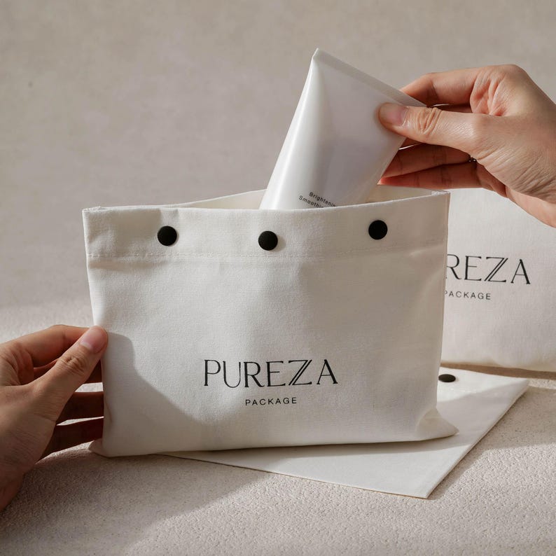 May include: A white canvas bag with the text "PUREZA PACKAGE" in black. The bag is open, and a white tube is being placed inside. The bag has three black snap closures at the top.