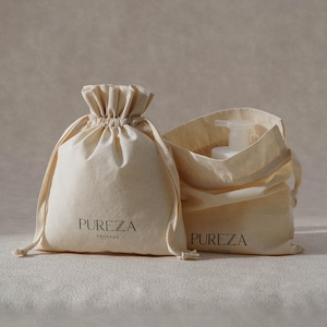 May include: Two beige drawstring bags with the text "PUREZA PACKAGE" printed in black. One bag is closed, and the other is open, revealing a white bottle with a pump dispenser. The bags are made of a natural fabric.