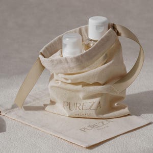 K&ouml;nnte beinhalten: Ein beigefarbener Stoffbeutel mit Kordelzug und einem Bandgriff, auf dem der Text PUREZA PACKAGE aufgedruckt ist. Im Beutel befinden sich zwei wei&szlig;e Kunststoffflaschen mit Pumpspendern. Ein passender flacher Stoffbeutel liegt darunter.