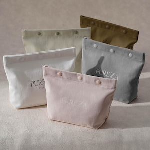 May include: A collection of five fabric pouches in pink, white, gray, cream, and olive green. Each pouch features a top flap with three snap buttons and the text PUREZA printed on the front. The pouches are arranged in a staggered formation.