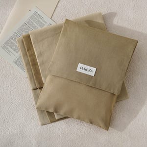 May include: A stack of folded, solid tan fabric pouches with a rectangular white label featuring the text PUREZA in black lettering centred on the front flap. The pouches are made of a smooth, matte material and are layered on top of each other.