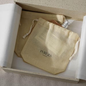 May include: A beige gift box contains two drawstring bags and a bottle. One bag has the text "PUREZA PACKAGE" printed on it. The box is lined with white paper. The bottle has a silver cap. The bags are made of a light-coloured fabric.