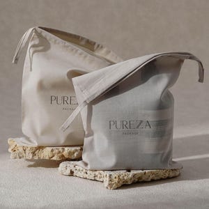 May include: Two beige drawstring bags, each with "PUREZA PACKAGE" printed on them. They rest on a textured, light-coloured stone surface. One bag is open, showing its contents.