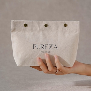 May include: A beige canvas pouch featuring three dark metal snap buttons along the top edge. The text PUREZA PACKAGE is printed in black in the center of the pouch. The fabric has a visible woven texture and a rectangular shape with rounded corners.