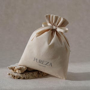 May include: A cream-coloured fabric bag with a drawstring closure and a tied ribbon. The bag is printed with the word "PUREZA" and "PACKAGE" in black. The bag rests on a light-coloured, textured stone surface.
