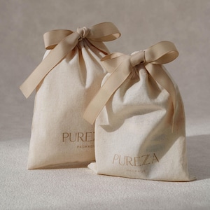 K&ouml;nnte beinhalten: Zwei cremefarbene Stoffbeutel mit Kordelzug und beigen Schleifen am oberen Ende. Jeder Beutel ist auf der Vorderseite mit dem Text PUREZA PACKAGE in dunkelbrauner Schrift bedruckt. Die Beutel bestehen aus einem naturfarbenen Textilmaterial.