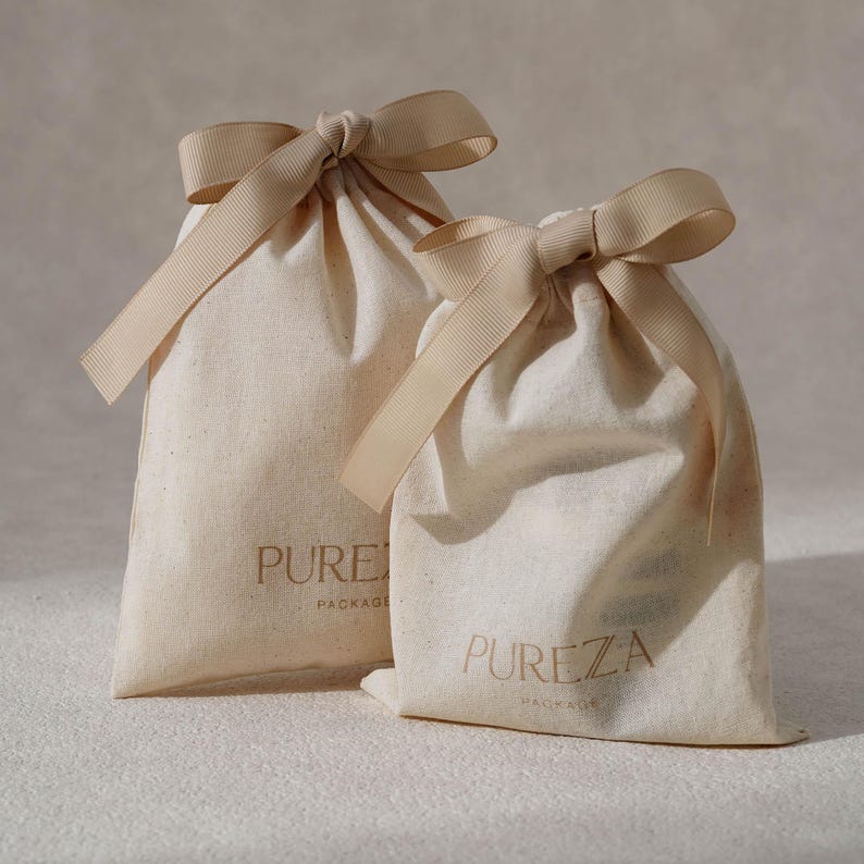 K&ouml;nnte beinhalten: Zwei cremefarbene Stoffbeutel mit Kordelzug und beigen Schleifen am oberen Ende. Jeder Beutel ist auf der Vorderseite mit dem Text PUREZA PACKAGE in dunkelbrauner Schrift bedruckt. Die Beutel bestehen aus einem naturfarbenen Textilmaterial.