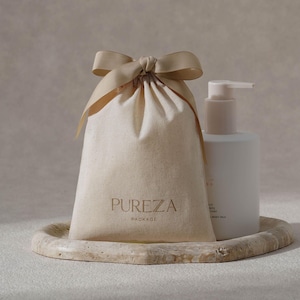 May include: A beige canvas bag with a tied ribbon, a white bottle with a pump, and a marble tray. The bag has the word "PUREZA" printed on it. The bottle has text on it. The items are arranged on a marble tray.