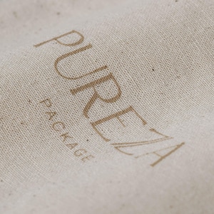 K&ouml;nnte beinhalten: Eine beige, texturierte Stoffoberfl&auml;che mit dem Wort PUREZA, das in einer gro&szlig;en, braunen serifenlosen Schrift gedruckt ist, wobei das Wort PACKAGE in einer kleineren, braunen Schrift direkt unter dem Buchstaben P steht.