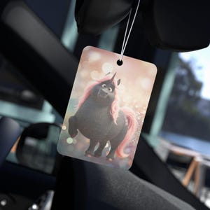 Könnte beinhalten: Ein rechteckiger Auto-Lufterfrischer mit einem Cartoon-Einhorn mit schwarzem Körper und rosa Mähne und Schweif. Der Lufterfrischer hängt an einer Schnur, mit einem verschwommenen Hintergrund aus Bokeh-Lichtern.