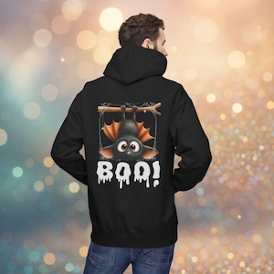 Könnte beinhalten: Schwarzer Kapuzenpullover mit einem Cartoon-Fledermaus-Motiv. Die Fledermaus hängt kopfüber an einem Ast, mit orangefarbenen Flügeln und großen Augen. Das Wort "BOO!" ist darunter in Weiß gedruckt.