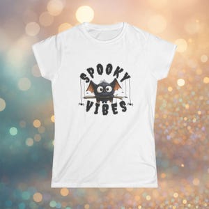 Könnte beinhalten: Weißes T-Shirt mit einem Cartoon-Fledermaus-Design. Die Fledermaus hat große Augen und Flügel und sitzt auf einem Holzstab. Der Text "SPOOKY VIBES" befindet sich über und unter der Fledermaus.