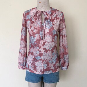 May include: A long-sleeved blouse with a floral print in shades of pink, blue, and white. The blouse has a tie at the neck and a gathered neckline.