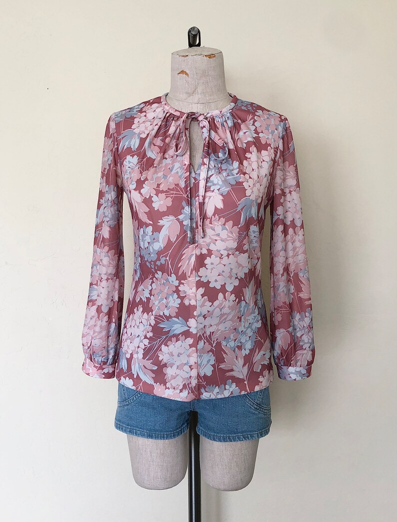 May include: A long-sleeved blouse with a floral print in shades of pink, blue, and white. The blouse has a tie at the neck and a gathered neckline.