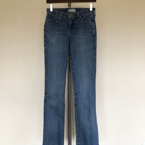 May include: Blue denim jeans hanging on a hanger. The jeans have a classic five-pocket design, a button and zipper closure, and a straight leg. The jeans are a medium wash denim.