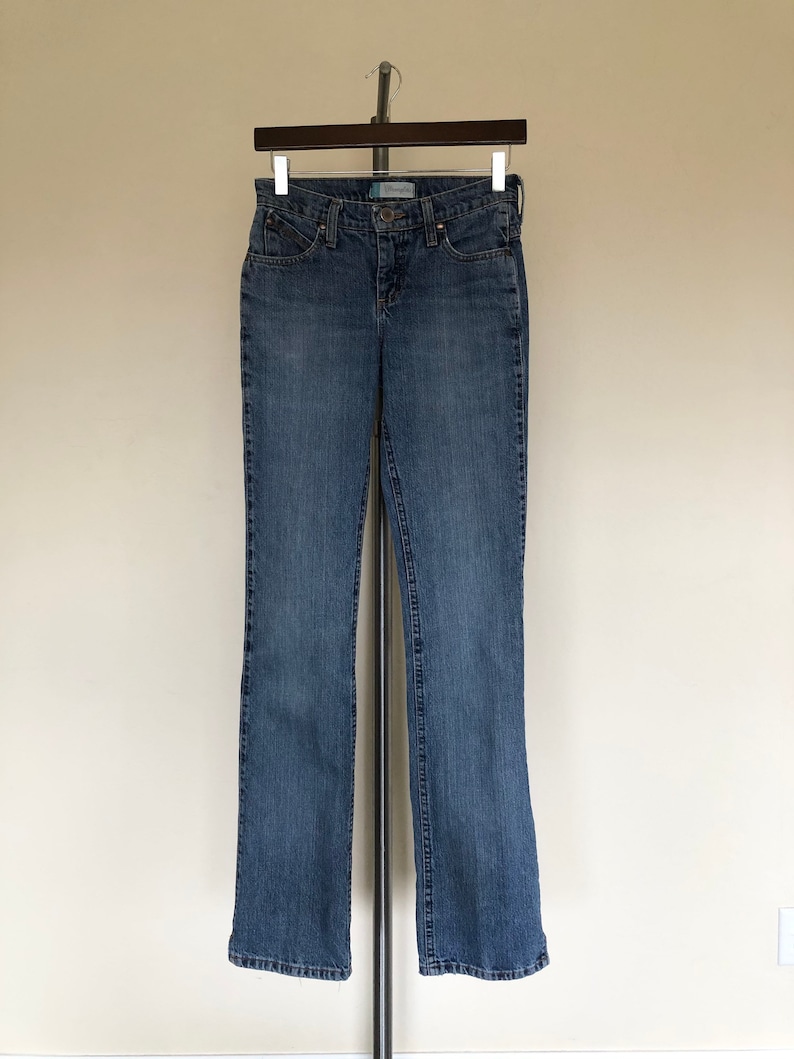 May include: Blue denim jeans hanging on a hanger. The jeans have a classic five-pocket design, a button and zipper closure, and a straight leg. The jeans are a medium wash denim.