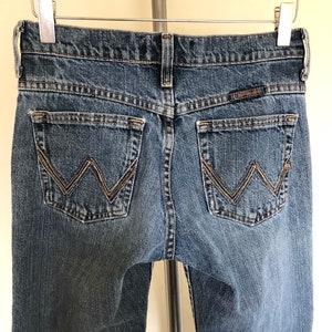 May include: Blue denim jeans with the Wrangler logo on the back pockets. The jeans are hanging on a metal hanger. The back pockets have a decorative stitching design. The jeans are a medium wash.