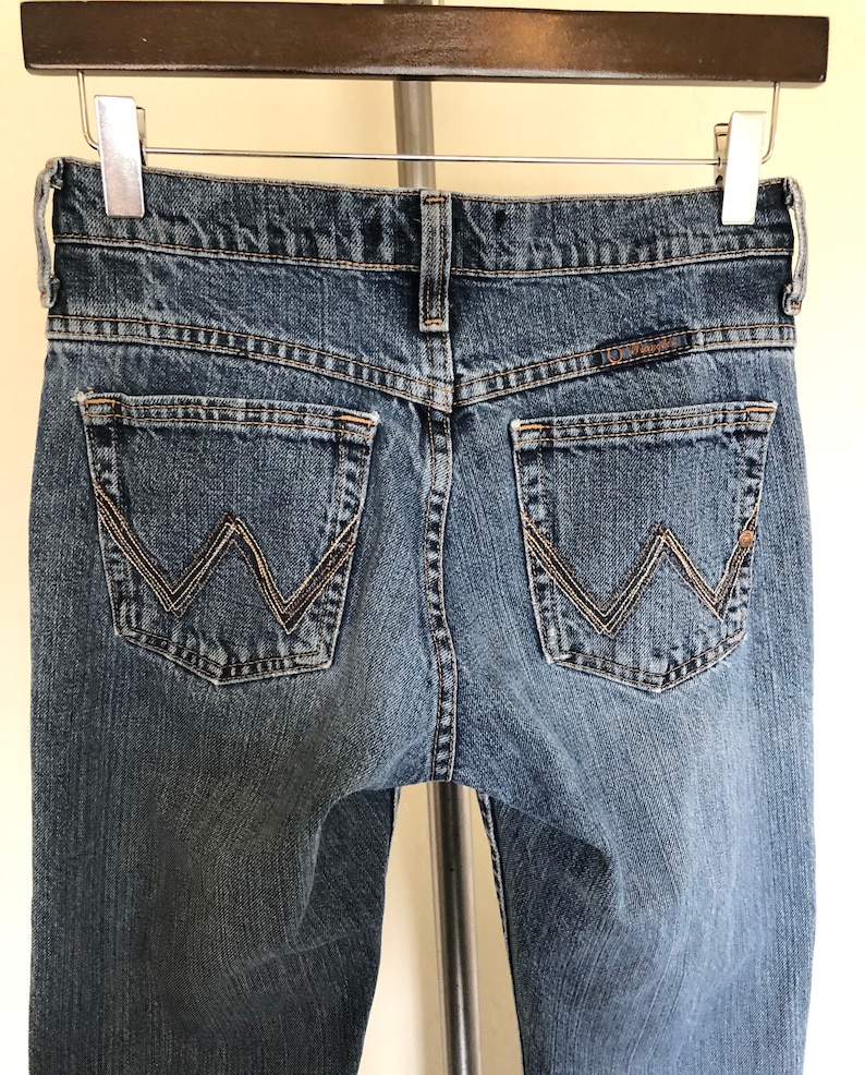 May include: Blue denim jeans with the Wrangler logo on the back pockets. The jeans are hanging on a metal hanger. The back pockets have a decorative stitching design. The jeans are a medium wash.
