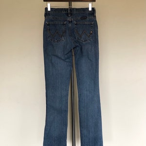 May include: Blue denim jeans with a classic five-pocket design. The jeans feature the Wrangler logo on the back pockets and a straight leg cut. The jeans are hanging on a metal hanger.