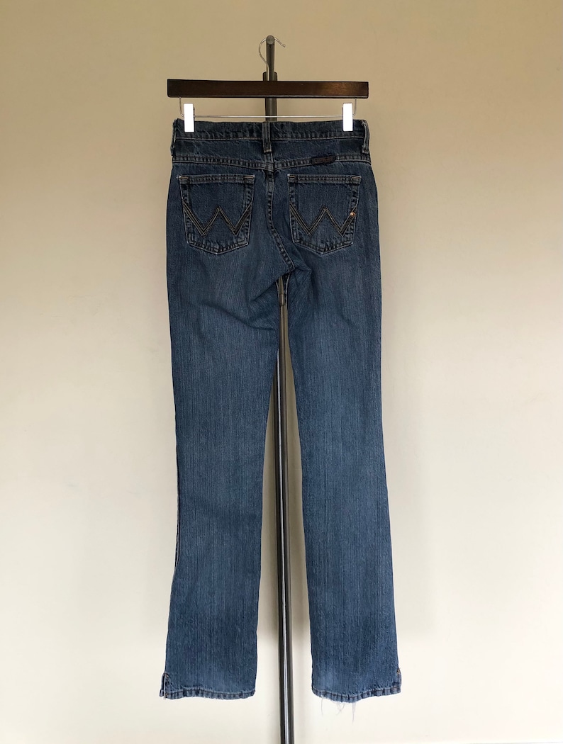 May include: Blue denim jeans with a classic five-pocket design. The jeans feature the Wrangler logo on the back pockets and a straight leg cut. The jeans are hanging on a metal hanger.