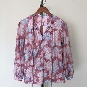 May include: A long-sleeved blouse with a floral print in shades of pink and blue. The blouse has a V-neckline and a tie at the neck.