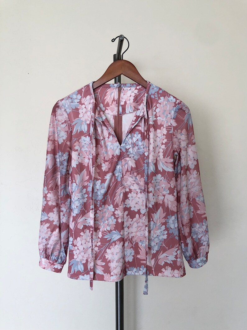 May include: A long-sleeved blouse with a floral print in shades of pink and blue. The blouse has a V-neckline and a tie at the neck.