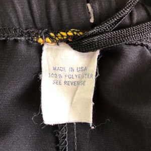 Op de afbeelding: Een wit label bevestigd aan een zwarte broek met een zwart trekkoord. Het label vermeldt "Made in USA 100% Polyester See Reverse."