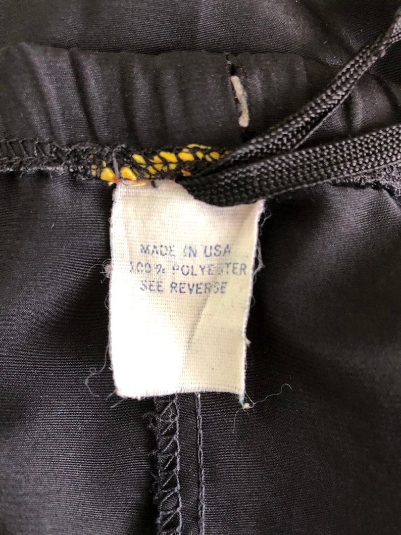Op de afbeelding: Een wit label bevestigd aan een zwarte broek met een zwart trekkoord. Het label vermeldt "Made in USA 100% Polyester See Reverse."