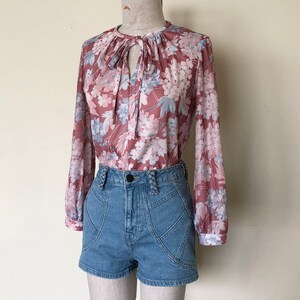 May include: A floral print blouse with a tie at the neck and long sleeves. The blouse is paired with denim shorts.