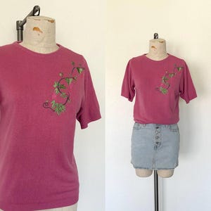 May include: A pink short-sleeved sweater with an embroidered floral design. The sweater has a crew neck and is made of a soft, lightweight material.