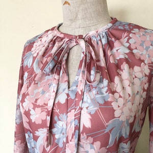 May include: A long-sleeved blouse with a floral print in shades of pink and blue. The blouse has a tie at the neck and a gathered neckline.