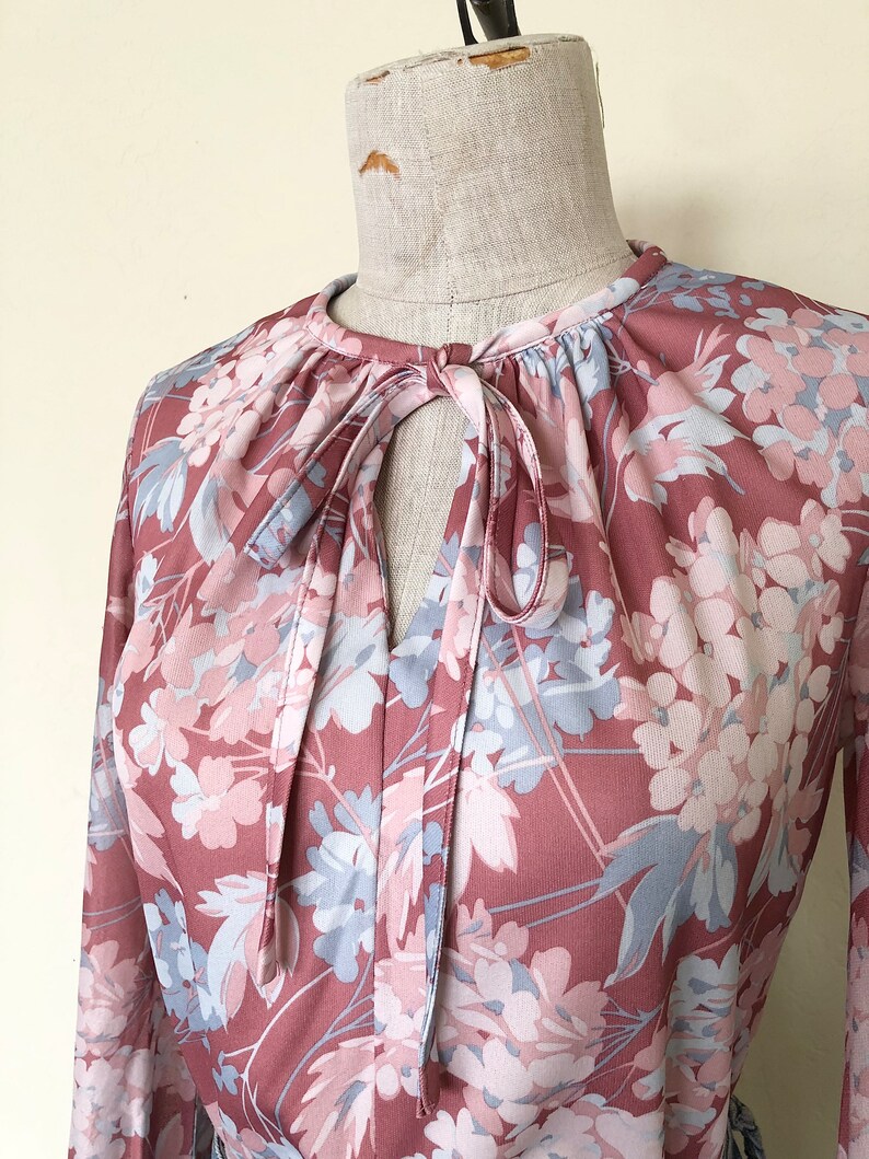 May include: A long-sleeved blouse with a floral print in shades of pink and blue. The blouse has a tie at the neck and a gathered neckline.