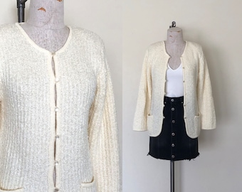 May include: A white knitted cardigan with a button-up front and two pockets. The cardigan is on a white mannequin.