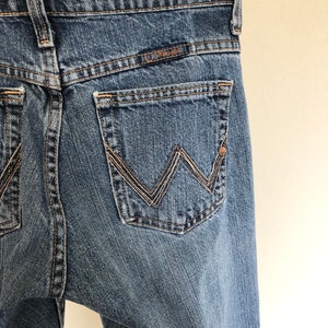 May include: Close-up of the back of blue denim jeans. The jeans feature a classic five-pocket design with a Wrangler logo above the back pocket. The stitching is a contrasting tan color, and the jeans have a worn appearance.