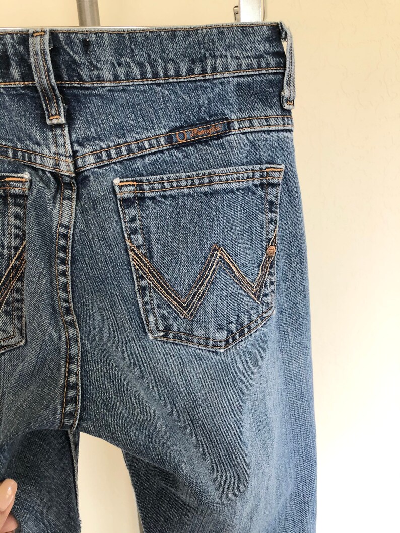 May include: Close-up of the back of blue denim jeans. The jeans feature a classic five-pocket design with a Wrangler logo above the back pocket. The stitching is a contrasting tan color, and the jeans have a worn appearance.