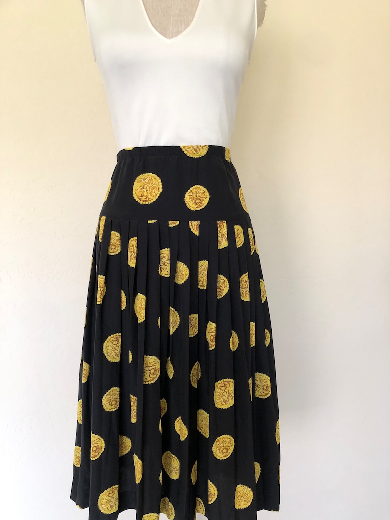 Op de afbeelding: Een zwarte geplooide rok met een gouden en bruine koekprint. De rok is midi-lang en heeft een aansluitende taille.