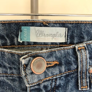 May include: Close-up of blue denim Wrangler Cowgirl Cut jeans. The jeans feature a button closure and a label that reads "Wrangler" and "Cowgirl Cut Jeans". The jeans are hanging on a metal bar.