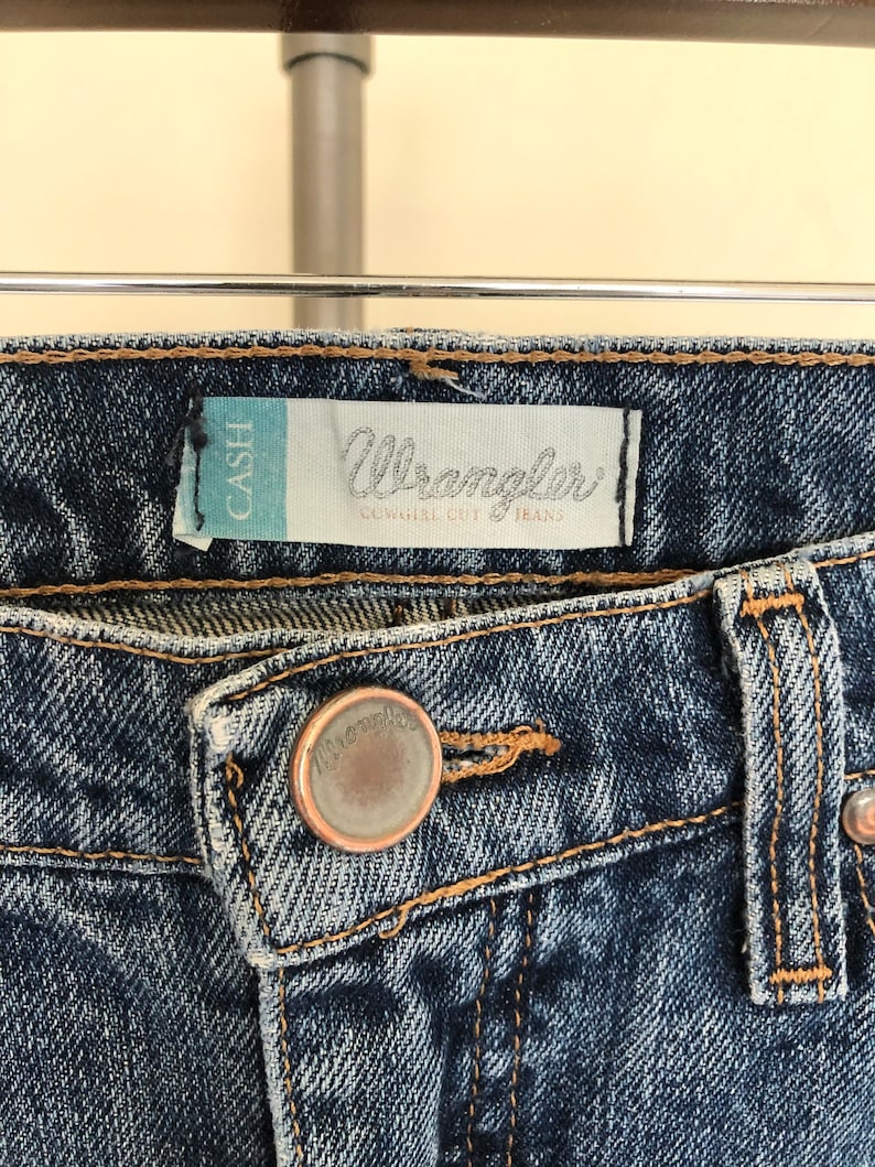 May include: Close-up of blue denim Wrangler Cowgirl Cut jeans. The jeans feature a button closure and a label that reads "Wrangler" and "Cowgirl Cut Jeans". The jeans are hanging on a metal bar.