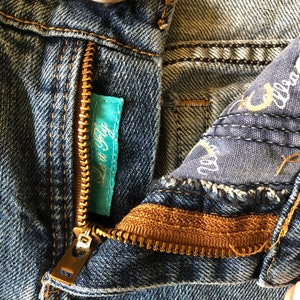 May include: Close-up of blue denim jeans with a partially open brass zipper. A small turquoise tag with the text "Lov It Fly" is visible. The jeans have a button closure and detailed stitching.