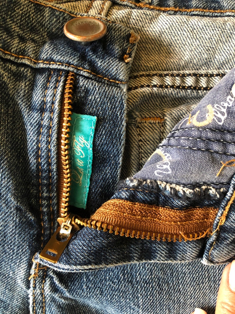 May include: Close-up of blue denim jeans with a partially open brass zipper. A small turquoise tag with the text "Lov It Fly" is visible. The jeans have a button closure and detailed stitching.