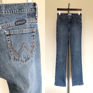 May include: Blue denim jeans with a classic five-pocket design. The jeans feature a straight leg cut and a Wrangler logo on the back pocket. The jeans are hanging on a wooden hanger.