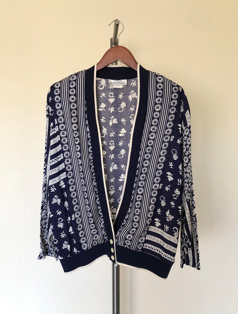 May include: A navy blue cardigan with a white floral pattern. The cardigan has a V-neck and a button closure. The label reads "Ellen Tracy".