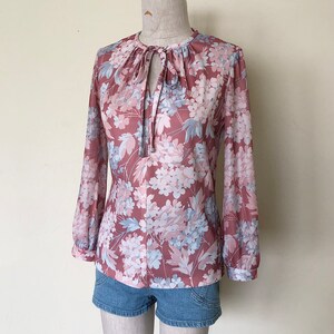 May include: A long-sleeved blouse with a floral print in shades of pink, blue, and white. The blouse has a tie at the neckline and a gathered detail at the shoulders.