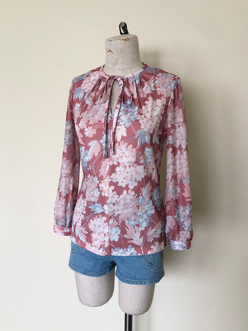 May include: A long-sleeved blouse with a floral print in shades of pink, blue, and white. The blouse has a tie at the neckline and a gathered detail at the shoulders.