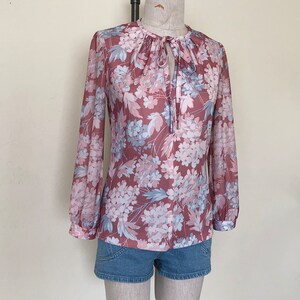 May include: A long-sleeved blouse with a floral print in shades of pink, blue, and white. The blouse has a tie at the neck and a loose fit.